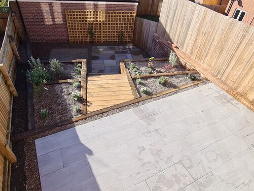 This image captures a charming garden layout with a wooden walkway leading to a trellis feature at the far end. The surrounding planting beds, filled with gravel and bordered by timber, host a variety of flowers and plants, creating a low-maintenance and visually appealing design. The natural wood tones and brickwork of the nearby structures complement the earthy aesthetic, making this space inviting and practical for outdoor enjoyment. The trellis may serve as a focal point, potentially supporting climbing plants in the future to enhance the greenery.