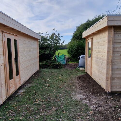 Two bespoke timber garden buildings supplied by Lugarde and designed and installed by Forevertimber. Both structures feature a natural timber finish with clean, minimalist designs. The building on the left includes a set of full-length glass-panelled double doors, while the building on the right has a simpler design with a single door and no visible windows. They are positioned on a grassy area with a clear space between them, offering flexibility for various uses such as storage, workshops, or recreational spaces. The surrounding greenery and open backdrop create a harmonious and practical garden setting.