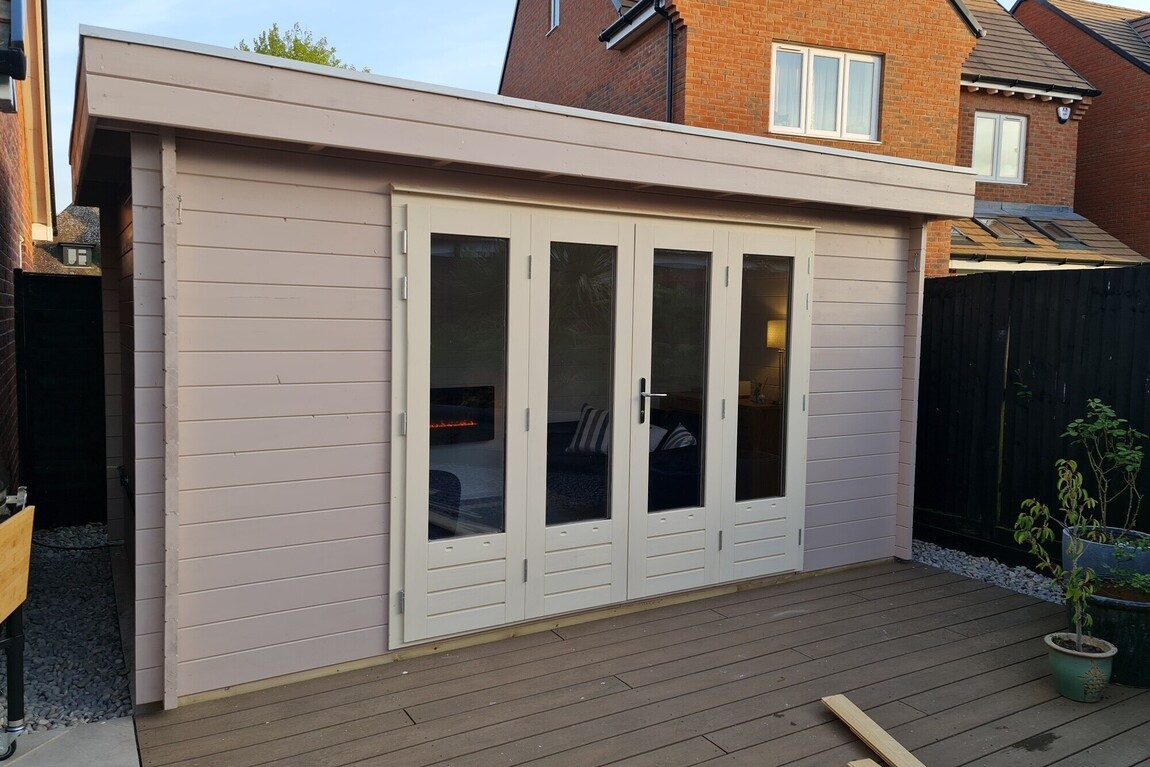 A bespoke timber garden building supplied by Lugarde and designed and installed by Forevertimber. This elegant structure features a soft pastel-colored exterior paired with white-framed full-length glass double doors. The doors and windows allow ample natural light to illuminate the interior, making it an inviting space. Set on a wooden decking area, the building is perfect for use as a home office, garden retreat, or additional living space. The clean, modern design complements the surrounding residential environment, blending functionality with style.