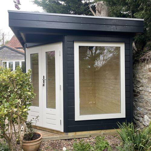 A stylish and modern timber garden building supplied by Lugarde and designed and installed by Forevertimber. This compact structure features a sleek black-painted exterior with white-framed glass-panelled double doors and a large rectangular window, allowing for plenty of natural light to fill the interior. The flat roof and clean lines give it a contemporary look, while the natural wooden interior adds warmth and versatility. Positioned against a stone wall and surrounded by greenery, this garden building is ideal for use as a home office, creative studio, or relaxing retreat. Its minimalist design integrates seamlessly into its natural surroundings.