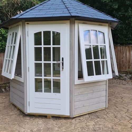 A charming hexagonal timber garden building supplied by Lugarde and designed and installed by Forevertimber. This elegant structure features a natural grey-stained wood exterior with white-framed glass double doors and matching windows. The windows are designed to open outward, allowing for excellent ventilation and a welcoming atmosphere. The building is topped with a classic black shingle roof, adding a touch of sophistication. Set on a patterned stone patio, this versatile garden house is ideal for use as a summerhouse, reading nook, or cosy retreat, blending functionality with timeless design.