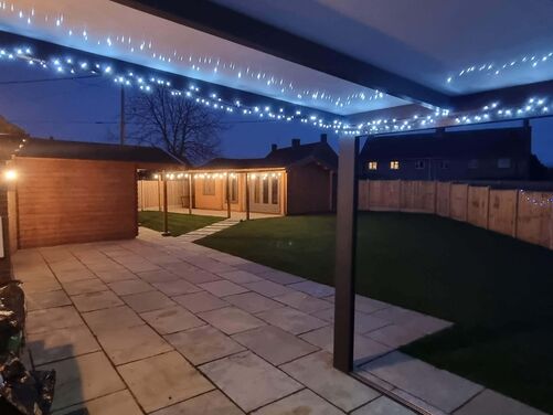 This image captures a cozy backyard at night, featuring a well-lit patio area and garden. String lights along the roofline of the patio create a warm and inviting ambiance, highlighting the modern, sheltered outdoor seating area. A glowing wall-mounted lamp on the brick wall adds to the soft illumination.

Beyond the patio, a pathway leads to a wooden garden structure with more string lights adorning its roof, offering a charming focal point in the space. The neatly trimmed lawn is enclosed by a wooden fence, providing privacy while maintaining an open and welcoming feel. This outdoor setting is perfect for evening relaxation or gatherings.