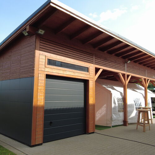 Modern timber garage and pavilion featuring a flat-sloped roof and a combination of sleek wood paneling and dark metal accents. The garage door is a stylish black panel design, complemented by a horizontal window for natural light. The pavilion area, supported by timber posts, includes an open-sided space ideal for hosting outdoor events or additional storage. Adjacent to the structure, a tented extension adds versatility for larger gatherings. The paved surroundings enhance functionality and accessibility. Designed and installed by Forever Timber, this building seamlessly integrates contemporary design with practicality and expert craftsmanship.