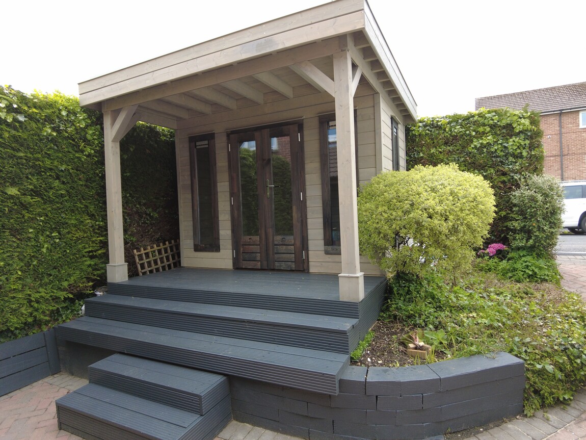 A modern wooden garden room designed and installed by Forever Timber, supplied by Lugarde. It features a covered porch, sleek grey decking, and large glass doors framed by wooden panels, creating a stylish and functional outdoor space. The surrounding landscaped garden adds a natural touch.