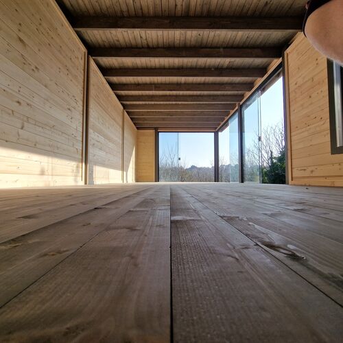 The interior of a bespoke timber garden building supplied by Lugarde and designed and installed by Forevertimber. This space features natural wood paneling on the walls, ceiling, and flooring, creating a warm and inviting ambiance. The ceiling has exposed beams, adding to the rustic yet modern aesthetic. The far end of the room showcases large glass panels that allow an abundance of natural light to fill the space, offering picturesque views of the surrounding outdoors. This versatile interior is perfect for a garden studio, workspace, or relaxation area.
