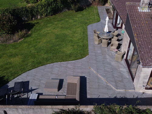 This image captures a beautifully landscaped outdoor area featuring a spacious patio that seamlessly connects to a well-maintained lawn. The curved edges of the patio design add a modern and elegant touch, while the choice of paving stones creates a sleek, cohesive look.

The seating areas are thoughtfully arranged, with a circular table surrounded by woven chairs for dining and a more relaxed lounge area with comfortable rattan furniture for casual gatherings. The expansive lawn provides ample space for outdoor activities and blends harmoniously with the natural greenery that surrounds the property.

The design balances functionality with aesthetic appeal, making this an inviting and versatile outdoor living space.