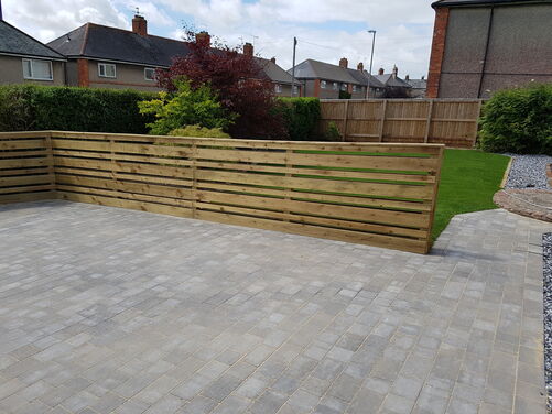 This backyard showcases a clean, modern design with a neatly paved patio area, a well-maintained lawn, and a sleek wooden privacy fence. The layout features a blend of functionality and aesthetics, with the fence providing separation between the paved section and the grassy area. The surrounding greenery adds a natural touch, while the gravel accents create a low-maintenance and polished appearance.