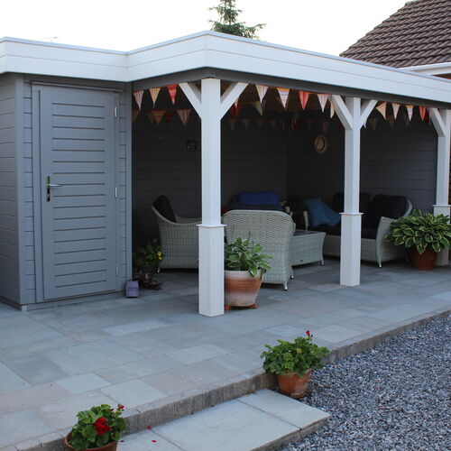 A bespoke garden gazebo supplied by Lugarde and designed and installed by Forevertimber. This stylish and functional structure features a combination of an open-sided seating area and an enclosed storage room with a grey-painted door. The gazebo's exterior showcases a grey finish with white posts and decorative cross-bracing, creating a modern yet inviting aesthetic. The spacious open area is furnished with wicker seating and potted plants, making it perfect for relaxing or entertaining. The paved flooring and bunting decorations add a welcoming touch, while the structure integrates seamlessly into the garden with its clean and contemporary design.