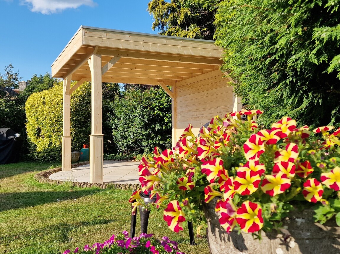 A bespoke timber garden gazebo supplied by Lugarde and designed and installed by Forevertimber. This open-sided structure features a natural wood finish and a sleek, flat roof supported by sturdy wooden posts with decorative bracing. The back wall provides added privacy and shelter, while the open layout creates an inviting space for outdoor relaxation or entertaining. Set on a circular paved patio and surrounded by lush greenery, it integrates beautifully into the garden. Vibrant red and yellow flowers in the foreground add a burst of colour to the serene outdoor setting.