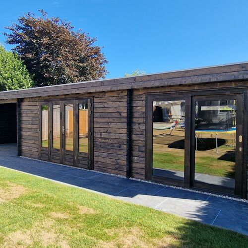 A bespoke timber garden building supplied by Lugarde and designed and installed by Forevertimber. This structure features a sleek, modern aesthetic with a dark-stained timber exterior, complementing the surrounding greenery. It includes multiple full-length glass doors and large windows, providing a bright and airy interior with excellent views of the garden. The flat roof and clean lines add a contemporary touch, while the stone-tiled pathway and patio enhance functionality and style. This spacious building is ideal for use as a multi-purpose garden retreat, office, or entertainment space.