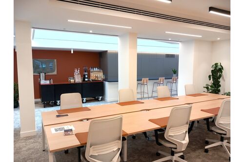 A modern office meeting room with long wooden tables and white chairs, featuring a coffee station in the background with an espresso machine, grinder, and supplies, set against a warm-toned wall with bar seating nearby.