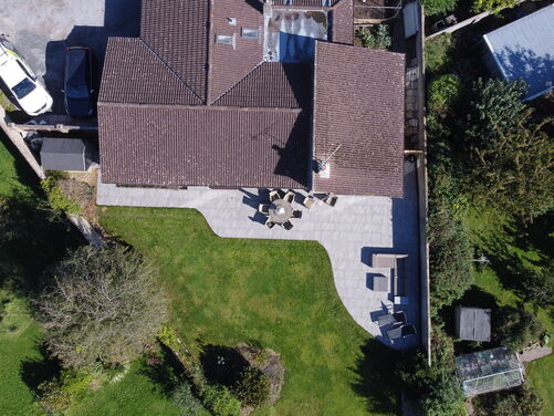 This aerial view showcases a well-maintained property with a spacious roof and thoughtfully designed outdoor spaces. The curved patio area blends seamlessly with the lush green lawn, creating a functional and aesthetically pleasing environment. The outdoor seating arrangement is positioned to take advantage of the expansive area, making it ideal for entertaining and relaxation.

The surrounding landscape includes trees and greenery, adding privacy and a natural touch to the property. The driveway area provides ample parking space, while the additional structures, like a greenhouse or shed, enhance the practicality of the space. The layout reflects a balance between landscaped gardens and functional areas, making it both inviting and practical.