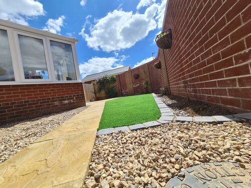This image displays a clean and well-maintained outdoor space featuring a combination of functional and aesthetic elements. The artificial grass lawn is neatly framed with grey brick edging, providing a vibrant green accent that contrasts beautifully with the warm tones of the brick walls and the golden paving slabs.

The surrounding gravel areas add texture and drainage functionality, while the brick wall adorned with hanging planters filled with colorful flowers adds a vertical dimension and charm to the space. The conservatory on the left, with its large windows, enhances the indoor-outdoor connection, making this area an inviting extension of the home, perfect for relaxation or light gardening. The overall design exudes simplicity, low maintenance, and an eye for detail.