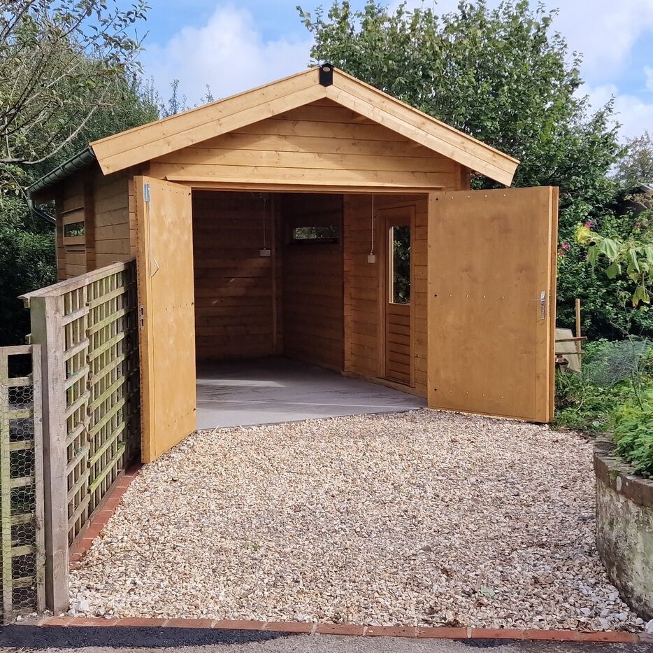 High-quality timber garage with a classic pitched roof, featuring wide double doors for easy access. The garage sits on a sturdy concrete base, surrounded by a neatly laid gravel driveway edged with brick. Natural wood tones blend seamlessly with the outdoor setting, offering a functional and aesthetically pleasing storage solution. This bespoke structure was supplied by Lugarde and meticulously designed and installed by Forever Timber, showcasing exceptional craftsmanship and attention to detail