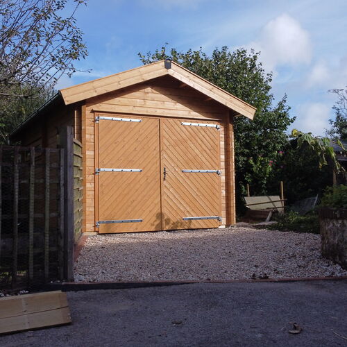 "Stunning timber garage with a classic pitched roof, constructed from high-quality natural wood. The design features wide double doors with a diagonal chevron pattern, secured with sturdy metal hinges and a central lock for enhanced functionality and security. The garage is set on a gravel driveway edged with bricks, blending seamlessly into the surrounding greenery for a natural and welcoming aesthetic. Supplied by Lugarde and expertly designed and installed by Forever Timber, this garage combines durability, practicality, and timeless charm."