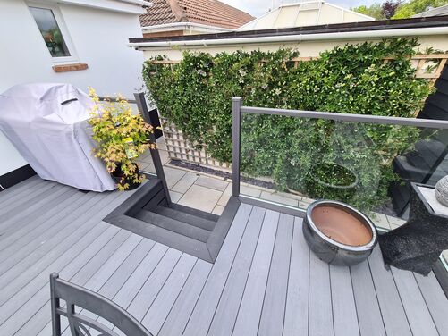 This space exhibits a sleek and contemporary deck design, featuring composite decking in a neutral gray tone that contrasts well with the surrounding greenery and white exterior wall. The addition of a glass balustrade not only ensures safety but also maintains an open and airy feel, allowing an unobstructed view of the garden area below.

The use of vertical planting on the trellis creates a lush green wall, adding texture and life to the scene. Functional elements like the built-in steps and outdoor furniture make the area both practical and stylish. The compact layout is ideal for small gatherings, outdoor dining, or simply enjoying some fresh air in a private, modern setting.