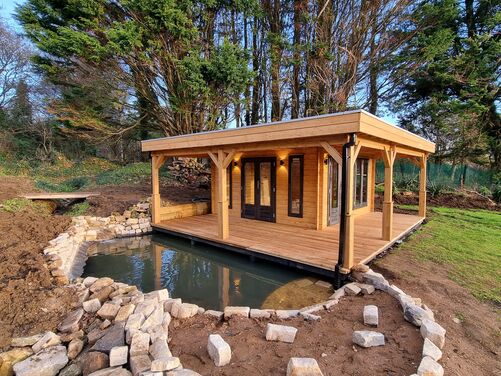 Beautiful timber garden room with a spacious covered decking area, overlooking a serene pond edged with natural stone. Nestled amidst lush greenery, this versatile outdoor structure offers a perfect retreat for relaxation or entertaining in a peaceful, natural setting