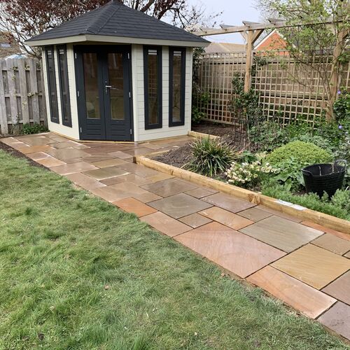 A bespoke hexagonal timber garden building supplied by Lugarde and designed and installed by Forevertimber. This charming structure features a cream-painted exterior with contrasting black-framed glass-panelled double doors and windows, offering a modern yet elegant aesthetic. The classic pitched roof is finished with dark shingles, adding a timeless touch. Set within a beautifully landscaped garden, the building is surrounded by a newly laid stone pathway and bordered flower beds, creating a tranquil and inviting space. This versatile garden building is perfect as a summerhouse, reading retreat, or hobby room, blending functionality with style.