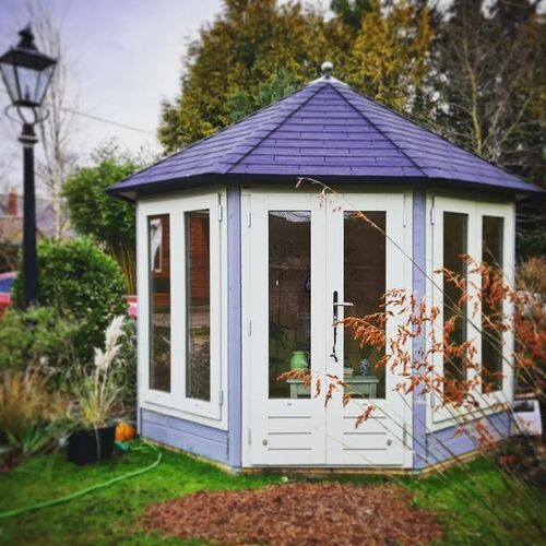 A charming hexagonal timber garden building supplied by Lugarde and designed and installed by Forevertimber. This elegant structure features a soft lavender-painted exterior with white-framed double doors and matching full-length windows on each panel, allowing plenty of natural light to brighten the interior. The building is topped with a slate-effect pitched roof, adding a classic and timeless aesthetic. Positioned in a beautifully landscaped garden, it is surrounded by lush greenery and accented by a vintage-style lamppost, creating a picturesque setting. This garden building is ideal for use as a cosy retreat, tea room, or creative workspace.