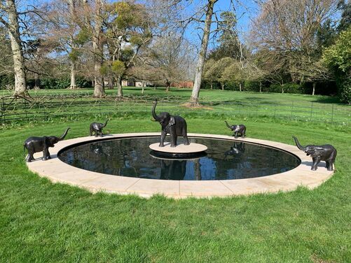 This image features a beautifully designed circular water feature set within a lush green lawn. The centerpiece includes a collection of elegant elephant sculptures, with the largest one positioned on a raised platform in the center, surrounded by smaller elephant figures on the edge of the water. The backdrop includes a serene landscape of tall trees and expansive grass, providing a tranquil and natural setting.
