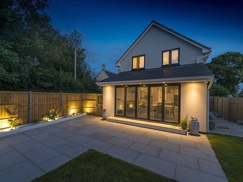This is a beautifully lit outdoor space that highlights the modern and clean design of the property. The combination of uplighting along the fence and the soft glow from the house's external lighting creates an inviting ambiance, particularly for evening gatherings. 

The large patio area is spacious and well-suited for outdoor furniture or dining arrangements, providing a functional and aesthetically pleasing extension to the indoor living space. The sleek bi-fold doors offer a seamless transition between the indoors and outdoors, enhancing the open-plan feel of the property. The neat fencing and landscaping add a sense of privacy and completeness to the area. This setup is ideal for relaxing or entertaining, day or night.