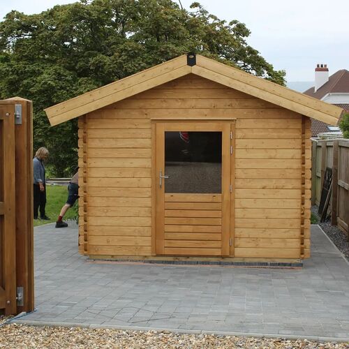 Sturdy timber garden building with a traditional pitched roof, crafted from natural wood for a timeless and rustic appearance. The front features a single door with a glass panel, allowing light into the interior while maintaining privacy. Set on a neatly laid paved surface, the building is surrounded by a well-maintained outdoor space with fencing for added privacy. Designed and installed by Forever Timber, this versatile structure is ideal for use as a garden shed, workshop, or recreational space, combining functionality with high-quality craftsmanship.
