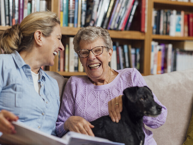 Carer and older woman laughing together at home with a small dog, reflecting warm, people-first care.
