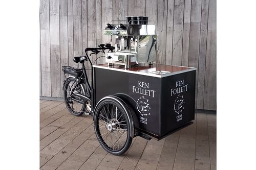 A three-wheeled cargo bicycle converted into a mobile coffee cart, featuring a stainless steel espresso machine on top and black panels branded “Ken Follett – Circle of Days,” set against a wooden wall background.