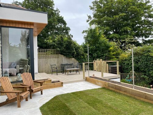 This image showcases a modern outdoor living space featuring a sleek extension with large glass panels that provide a seamless connection between the indoors and outdoors. The backyard includes a decked seating area with a mix of contemporary and rustic furniture, surrounded by lush greenery for privacy. The lawn and raised planters add natural elements, while the clean layout ensures functionality.

The wooden Adirondack chairs and neutral tones enhance the relaxed and inviting atmosphere.