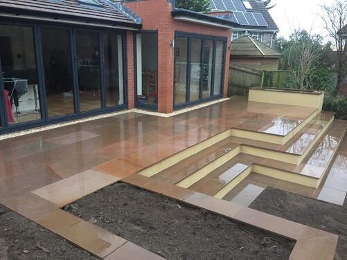 This image features a modern outdoor patio area with large, polished stone tiles that give a sleek and reflective finish, especially noticeable under wet conditions. The multi-leveled steps provide a dynamic layout, complementing the contemporary design of the house with its large glass doors and red brick exterior. The steps seem to lead into a lower garden area, creating a connection between the patio and the surrounding natural space. This setup is ideal for a seamless indoor-outdoor living experience.