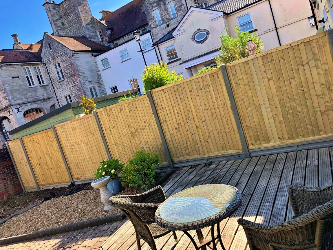 Charming backyard featuring a timber decked seating area with wicker furniture, surrounded by a neat wooden panel fence for privacy. The backdrop of historic architecture adds a unique character, blending modern outdoor living with timeless elegance.