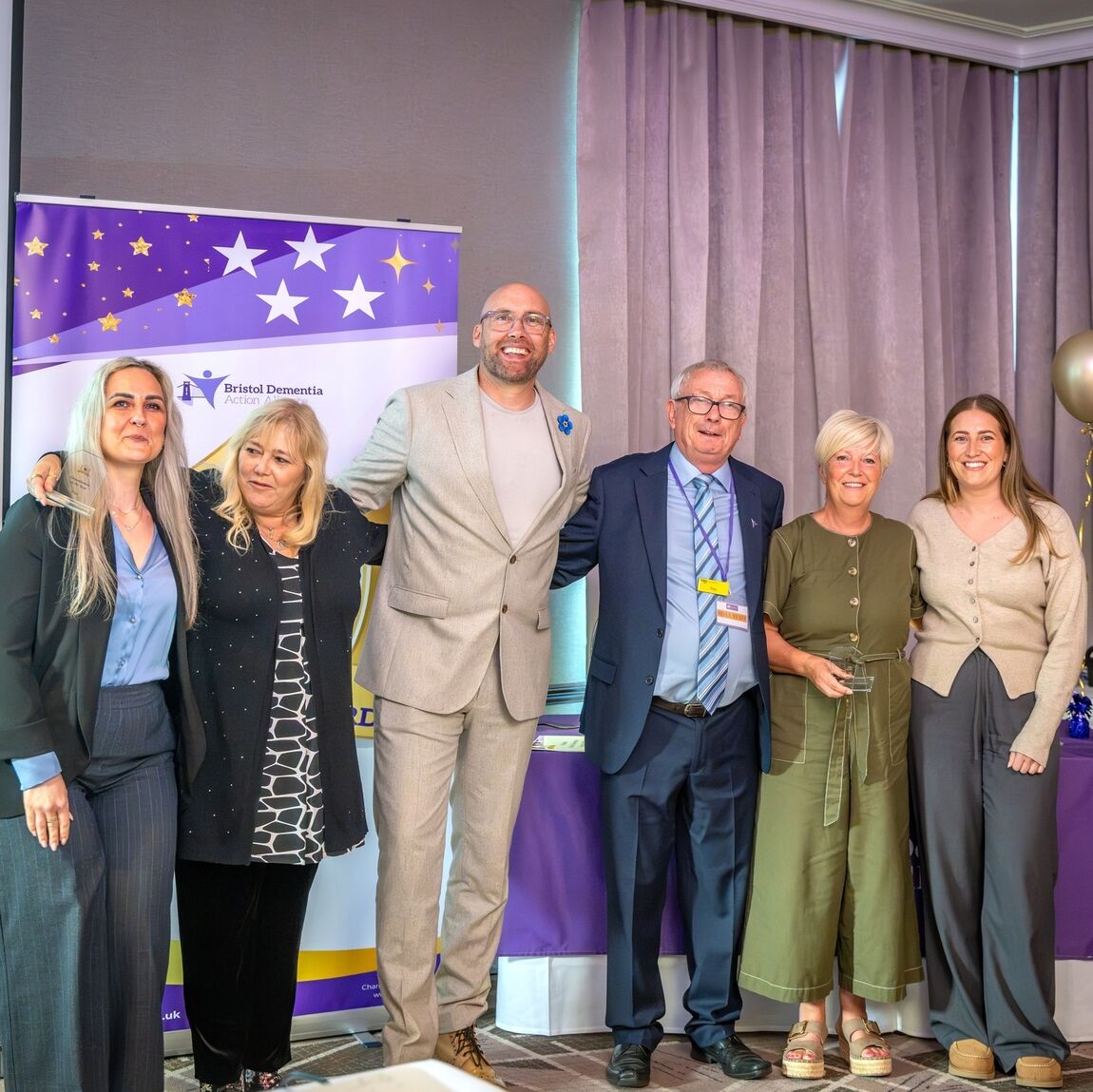 Six attendees pose with glass trophies in front of a Bristol Dementia Action Alliance banner at the 2026 awards.