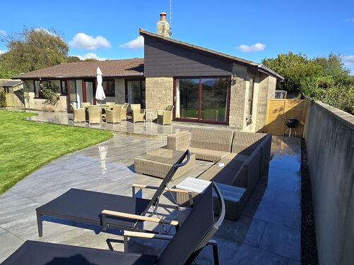 The image showcases a beautifully designed outdoor space that combines a large, polished gray tiled patio with a lush, well-maintained lawn. The patio features a variety of seating options, including wicker chairs arranged around a table with an umbrella for shade and a comfortable sectional sofa for lounging. Sun loungers add a touch of luxury, perfect for relaxation. The house, with its stone and wood exterior and large glass windows, opens directly onto this outdoor area. A low wall and mature landscaping, including shrubs and trees, provide privacy and natural beauty. The overall design emphasizes comfort, functionality, and a seamless connection between the indoor and outdoor spaces.