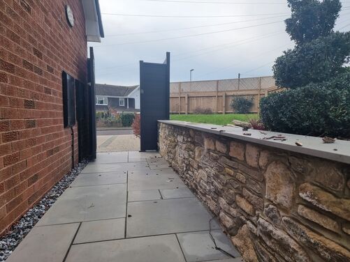 The image depicts a neat and stylish outdoor pathway alongside a brick house. The pathway is paved with light gray stone slabs, bordered on one side by a stone wall with natural stones and a smooth coping stone on top. On the opposite side, a narrow strip of decorative gravel runs along the base of the brick wall.

At the end of the pathway, a modern black wooden gate leads to a driveway or outdoor area. In the background, a lawn and wooden fence with a trellis top are visible, providing privacy and a natural touch. The space is well-organized, with a combination of traditional and modern elements, creating a clean and appealing look.