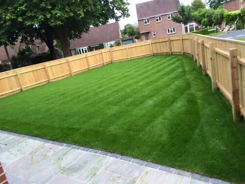 The image shows a well-maintained garden featuring a neatly mowed lawn with visible striping, giving it a professional and polished look. The lawn is surrounded by a sturdy wooden fence, providing both privacy and a clear boundary for the space. The area is bordered by a paved pathway made of light-colored stone slabs, creating a clean and tidy transition between the grass and any adjacent areas. In the background, trees and neighboring houses enhance the suburban and tranquil atmosphere of the setting.