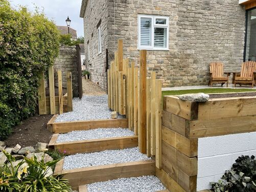 The image shows a neatly landscaped outdoor pathway and staircase made of wooden sleepers and filled with light gray gravel. The stairs connect different levels of the garden, bordered by wooden posts that add a modern, rustic charm. The surrounding area includes planted greenery, such as flowering shrubs and decorative plants, which soften the structured design. In the background, the stone facade of the house and a set of wooden Adirondack chairs are visible, placed on a paved patio adjacent to a small lawn. The setting combines natural materials and clean lines, creating a well-organized and visually appealing garden space.