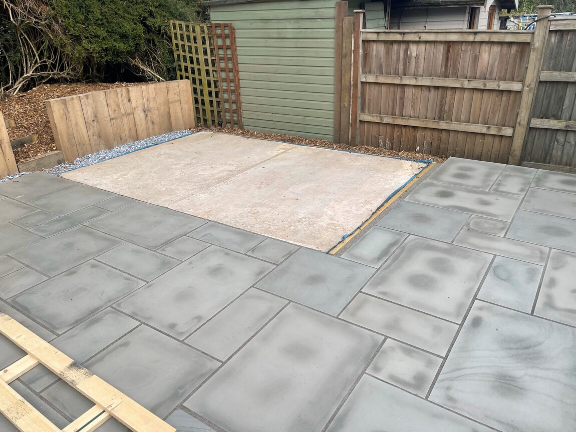 Prepared garden area featuring a newly laid concrete base surrounded by freshly installed grey paving slabs. The space is bordered by wooden fencing and trellises, creating a private and defined outdoor setting. A gravel edging adds a neat finish to the boundary of the concrete base, which appears to be ready for the installation of a garden building or structure. This clean and functional layout is designed to enhance the utility and aesthetics of the outdoor area.