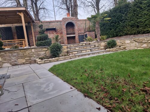 The image features a beautifully landscaped garden with a blend of stone, greenery, and well-crafted design elements. In the foreground, a paved pathway made of light gray stone slabs curves alongside a lush green lawn, scattered with autumn leaves. 

A stone retaining wall, constructed with natural stones, borders the lawn and leads up to a raised area via stone steps. The raised section features an outdoor seating area under a wooden pergola adorned with string lights, and a red brick structure with an arched opening serves as an outdoor fireplace or barbecue. Decorative spiral-shaped shrubs, a small palm tree, and other greenery add a touch of elegance and contrast to the hardscape. Tall hedges and mature trees in the background enhance privacy and provide a natural, serene backdrop.