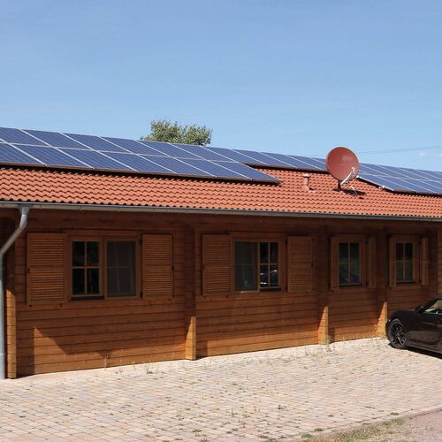 Eco-friendly single-story timber house with a red-tiled roof, equipped with solar panels for sustainable energy use. The exterior showcases warm-toned timber walls with decorative wooden shutters framing the windows, adding a rustic yet refined touch. A paved driveway extends along the front, providing ample parking space. Designed for modern living, the house incorporates environmentally conscious features while maintaining timeless aesthetic appeal. Supplied by Bertsch Holzbau and expertly designed and installed by Forever Timber, this timber home offers a perfect blend of sustainability, functionality, and craftsmanship.