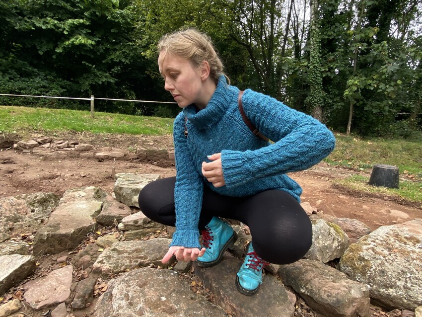 Lucy with plaited blonde hair, is wearing a turquoise roll-neck jumper, leggings and bright blue DR Marten boots. She is crouching and pouring an imaginary urn like Sabrina towards the ground. She is crouched on a low plinth on a Roman excavation site where a dedication to Sabrina would have stood.