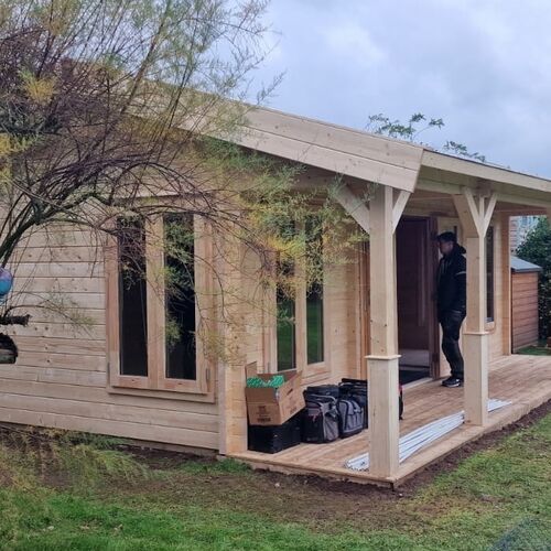 A bespoke timber garden building designed by Lugarde and crafted by Forevertimber. This structure features a rustic charm with its natural wood finish and sloped roof extending to form a covered veranda. The design includes large windows and a door with glass panels, allowing for ample natural light. The veranda adds a cozy outdoor seating area, sheltered by the roof supported by sturdy wooden beams. This versatile and charming building is ideal for a garden retreat, studio, or additional living space, blending harmoniously with its natural surroundings.