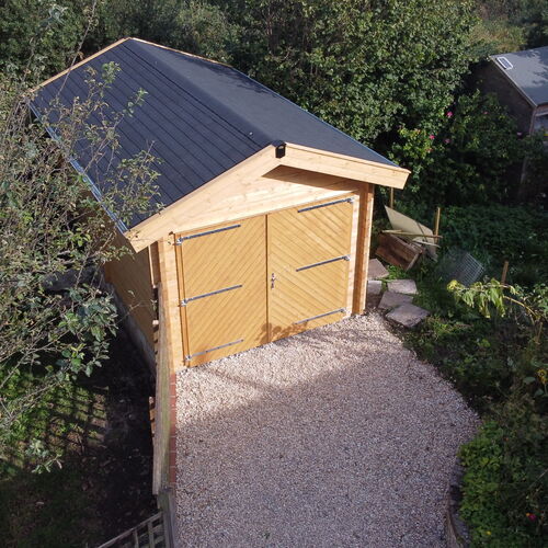 "Rustic timber garage with a pitched roof, constructed from premium-quality wood and complemented by double doors featuring cross-brace detailing and sturdy metal hinges. The roof is covered with durable dark roofing material for weather resistance, while the structure is nestled in a lush garden setting, surrounded by greenery. A well-maintained gravel driveway, bordered by bricks, leads to the garage, creating a natural and inviting entrance. Supplied by Lugarde and thoughtfully designed and installed by Forever Timber, this garage combines practicality with natural charm to seamlessly integrate into the landscape."