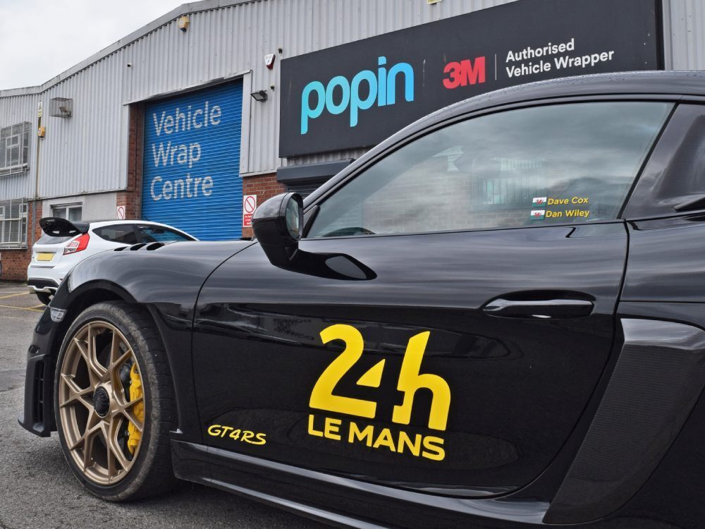 Yellow branded vehicle graphics on black company Porsche GT4 RS outside Popin in Cardiff.