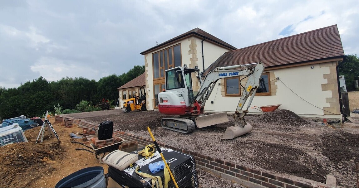 Construction site of a modern home featuring large windows and cream exterior walls with stone detailing. The groundwork is underway, with heavy machinery and building materials on-site, preparing for landscaping or additional outdoor structures. The spacious setting is surrounded by greenery, offering potential for a stunning finished project.