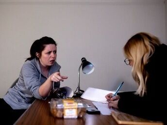 Karla is sitting on a chair wearing a grey hoody, glaring and pointing at Mags. Mags is hunched forward writing notes with the help of a small spot lamp. Her blonde hair is trailing down the side of her face and past her glasses.