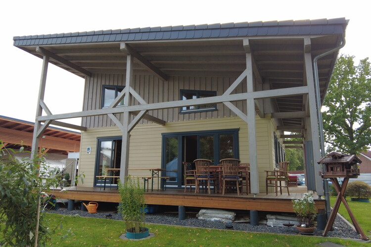 This modern accommodation is a two-story house with a black roof, white walls, and large windows. The front features a small porch adorned with potted plants. It is nestled in lush green lawns, among trees and colorful foliage, under a clear blue sky.
