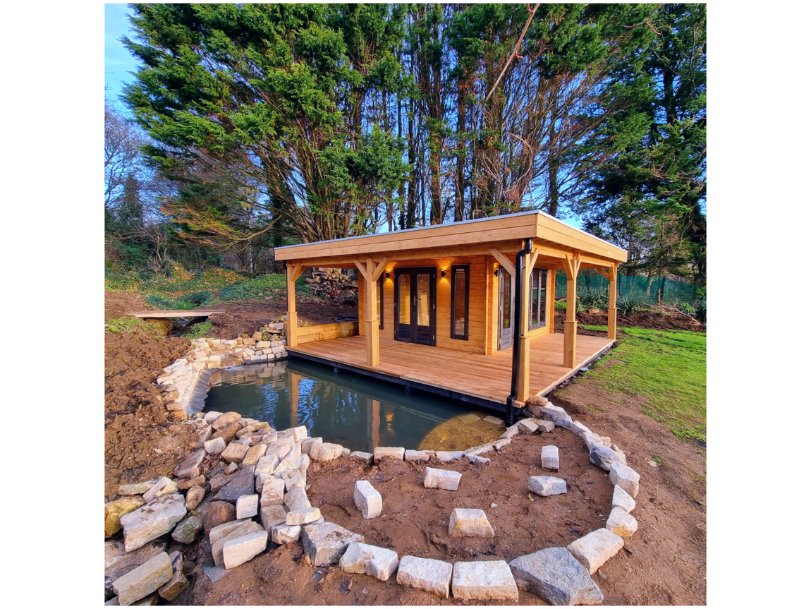Beautiful timber garden room with a spacious covered decking area, overlooking a serene pond edged with natural stone. Nestled amidst lush greenery, this versatile outdoor structure offers a perfect retreat for relaxation or entertaining in a peaceful, natural setting