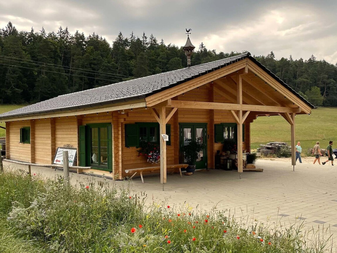 Rustic yet modern commercial timber building featuring a pitched roof with traditional wooden beams and shingles, creating a classic alpine-inspired aesthetic. The exterior showcases natural wood cladding with green-framed windows and doors, complemented by decorative shutters for a cozy and inviting look. A spacious covered entrance supported by sturdy timber posts provides a sheltered area, ideal for outdoor seating or product displays. Surrounded by lush countryside with wildflowers and open fields, this building seamlessly integrates into its natural environment. Designed and installed by Forever Timber and supplied by Bertsch HolzBau, this structure combines functionality, craftsmanship, and timeless charm, making it perfect for commercial use in scenic locations.