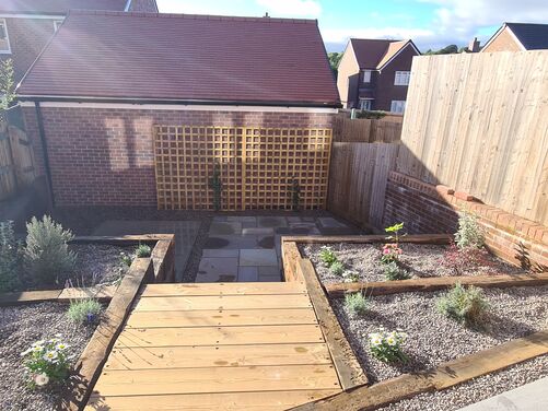 The image shows a compact and well-organized backyard space with a mix of natural and hardscaped features. A wooden pathway leads down to a lower area with gravel beds on both sides, bordered by wooden sleepers. These gravel beds are planted with small shrubs, flowers, and herbs, adding greenery and texture to the area. 

At the far end, a brick wall with lattice trellis panels provides a decorative and functional backdrop, potentially for climbing plants. The yard is enclosed by wooden fencing, offering privacy. The layout is simple yet functional, making good use of the available space while creating an inviting and low-maintenance garden.
