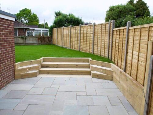 The image depicts a neat and minimalist outdoor garden area. The foreground features a light-colored paved patio with smooth, rectangular slabs, providing a clean and structured appearance. Steps made of wooden sleepers lead to a slightly elevated lawn area, bordered by wooden planters filled with white decorative stones. The lawn is surrounded by a tall wooden fence with concrete posts, offering privacy and a uniform look. The design balances functional outdoor space with simple landscaping, creating a tidy and inviting atmosphere.